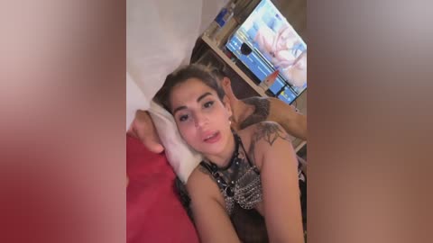 A video shows a young woman with light skin, short gray hair, and tattoos lying on a red bed, watching TV. She has a neutral expression.