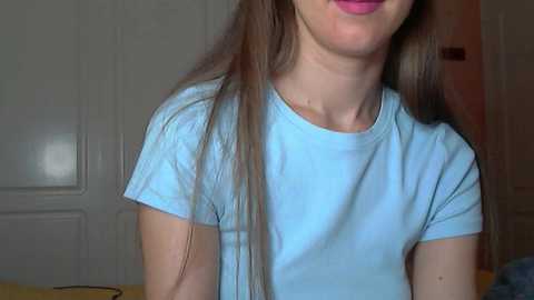 Video of a young woman with long brown hair, light skin, and a light blue t-shirt, smiling slightly. Background includes a white door and a dimly lit room.