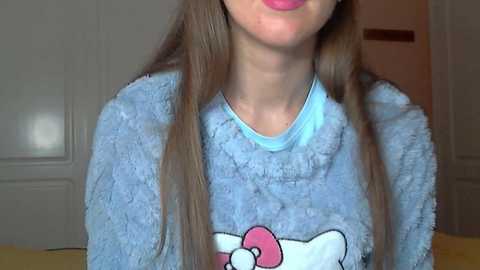 Video of a young woman with long brown hair, wearing a fluffy blue Hello Kitty pajama top. Her face is partially visible, showing a hint of pink lipstick. Background includes a white door and part of a closet.