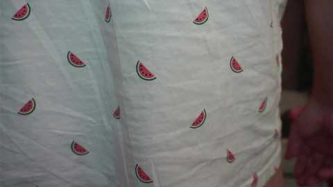 A close-up video of a person's lower torso, featuring white fabric with small, evenly spaced watermelon slices embroidered in green and red. The person's skin tone is visible in the background.
