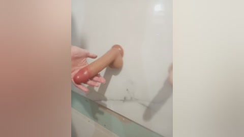 A video captures a hand holding a realistic, uncircumcised, tan-colored dildo against a mirror. The background shows a tiled bathroom wall with a greenish-blue and white pattern. The image is intimate and explicit.