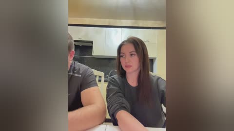 Media: A video shows a woman with long dark hair, wearing a black sweater, sitting at a kitchen counter, gazing at a man with short hair, wearing a gray shirt. The background features modern kitchen cabinets and a black stove.