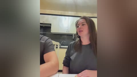 Media: A video of a woman with long brown hair in a grey top, smiling and holding a piece of paper, in a modern kitchen with light wood cabinets and black countertops.