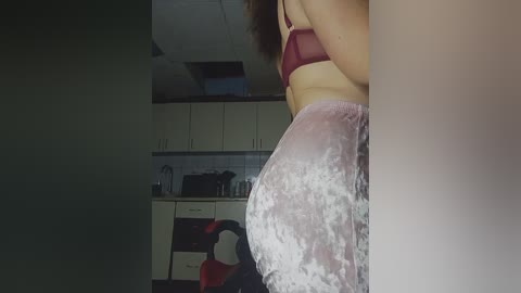 Media: Video of a woman with fair skin wearing a maroon bra and velvet pants, standing in a dimly lit kitchen with white cabinets and appliances.