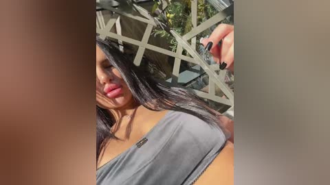 Media: Video of a woman with long, straight black hair, wearing a gray tank top, with a hand brushing her hair. Background features a metal fence and green foliage.