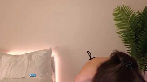 Video of a woman with long brown hair, wearing a black bra, lying on a bed with white pillows and a blue phone. Background features a green plant and beige wall.
