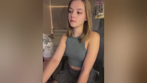 Media: Video of a young Caucasian woman with light skin and straight blonde hair, wearing a sleeveless gray top and gray pants, seated in a dimly lit bedroom with beige walls and a bed with white bedding.