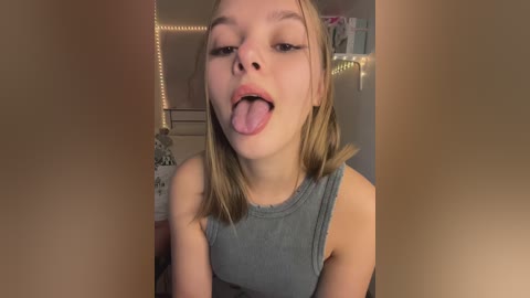 Media: Video of a young, fair-skinned, blonde-haired girl with a playful expression, sticking out her tongue and wearing a grey tank top, standing in a cozy, dimly-lit room with a string of fairy lights.