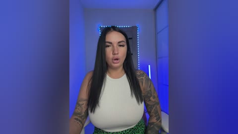 Video of a tattooed woman with long black hair, wearing a white tank top and green skirt, in a dimly lit hallway with blue LED strip lights.