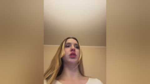 A video of a blonde woman with fair skin, wearing a white top, standing in a beige hallway. Her expression is neutral.