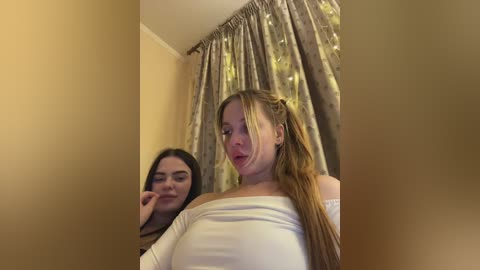 A video of two young women, one with blonde hair in a ponytail and another with long dark hair, standing in a room with beige walls and floral-patterned curtains.