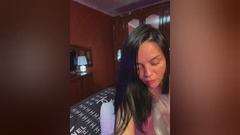 Video of a young Asian woman with straight black hair, wearing a white tank top, sitting on a bed with a patterned duvet, in a dimly lit, cozy bedroom.