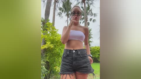 Media: Video of a slender, fair-skinned woman with brown hair tied back, wearing a white tube top and high-waisted, frayed denim shorts, standing outdoors against a backdrop of greenery and palm trees.