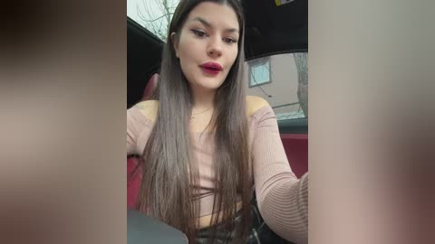 Media: Video of a young woman with long brown hair, wearing a pink off-shoulder top, seated in a red car, looking at her phone.