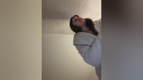 Media: A video of a young woman with long brown hair, wearing a white sweater, looking upward with a serene expression. The image features a plain, off-white ceiling and walls, creating a minimalist backdrop.