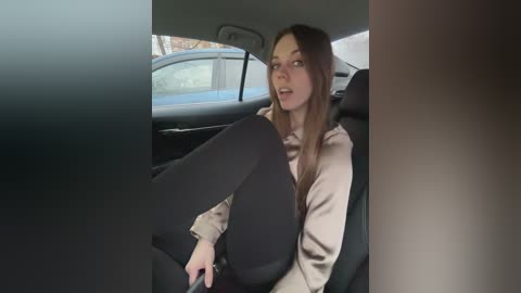 Media: Video of a young woman with light skin and long brown hair, wearing a beige jacket and black leggings, sitting in a car with the door open, legs out.