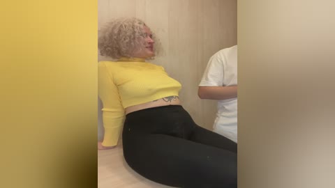 Media: A video showing a woman with curly gray hair, wearing a yellow turtleneck and black leggings, sitting on a light wooden floor. Another person, partially visible, stands beside her. The background is beige and yellow.