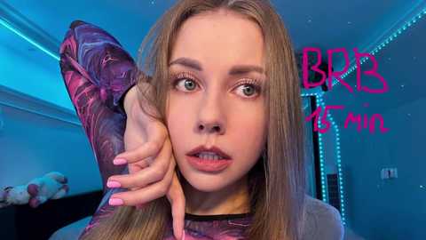 Media: Video of a young, fair-skinned woman with long, straight blonde hair, wearing a marbled purple long-sleeve top, looking surprised. Background features blue LED lights and the text \"BBB 2:1\" in pink.