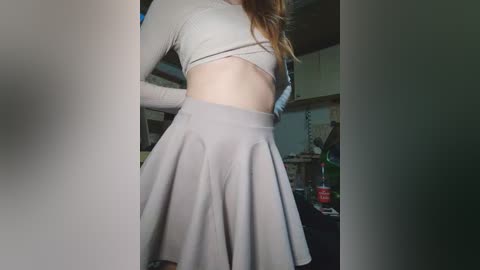 Video of a light-skinned woman with long brown hair, wearing a cropped white crop top and a high-waisted, pleated gray skirt, in a dimly lit kitchen.