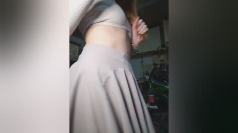 A video of a woman in a grey ribbed crop top and high-waisted pleated skirt, standing indoors near a cluttered bookshelf.