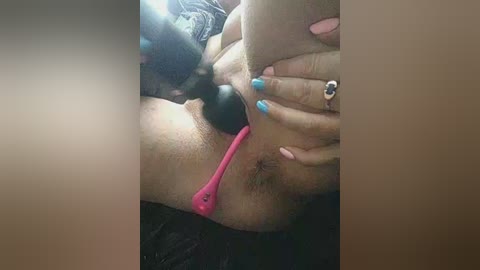 Video of a person with light brown skin and manicured nails, using a pink vibrator on their shaved pubic area. The background is blurred.