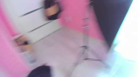 A blurred video captures a person with brown skin, wearing pink and black clothes, standing in a white-walled room with a reflective surface. The image is out of focus, creating a dreamy, surreal effect.