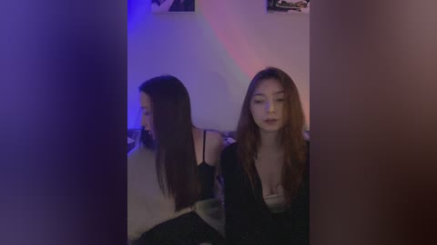 Media: Video of two Asian women with long hair, one in a black top, the other in a white top, sitting side by side, dimly lit room with blue and purple lights.