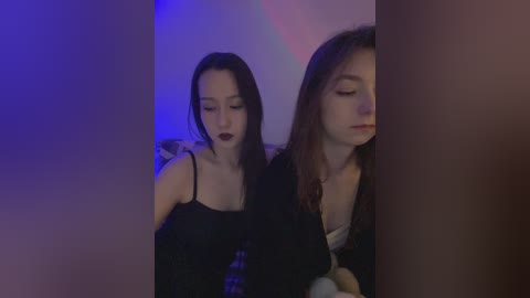 Media: Video of two young women with pale skin, one with dark hair and the other with brown hair, dressed in black, standing close together in a dimly lit room, with blue and pink light accents.