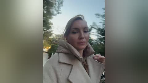 Video of a blonde woman with fair skin, wearing a beige coat, standing outdoors near trees and a blurred building.