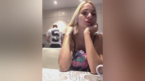 Media: Video of a blonde woman with medium-length hair, wearing a floral-patterned top, leaning on her hands at a tiled counter. In the background, a man sits on a white chair, facing away.