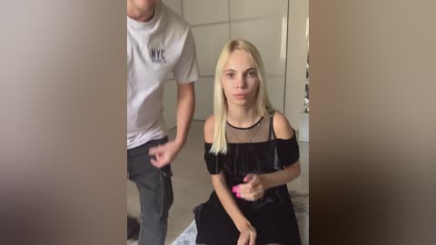 Media: Video of a blonde woman with long hair, wearing a black off-shoulder dress, sitting on a bed, looking upset. A man in a white T-shirt and jeans is in the background, appearing to be in motion.