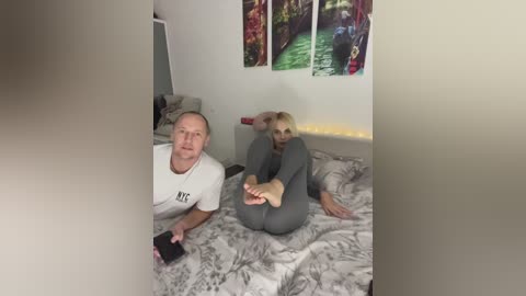 Media: Video of a middle-aged man with short hair and a woman with long blonde hair, both in gray pajamas, lying on a bed with a floral-patterned duvet.