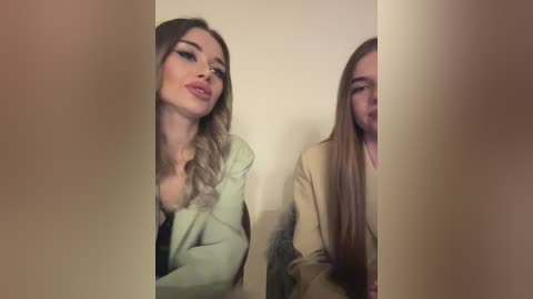 Media: Video of two young women with long hair, one with blonde ombre, the other with straight brown hair, both wearing blazers, smiling, partially obscured by a blurred, beige wall.