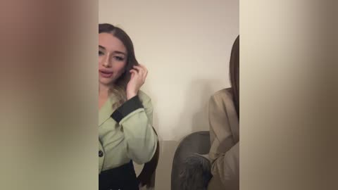 Media: A video captures a woman with light skin and straight, brown hair, wearing a green jacket, talking on a phone, partially obscured by a blurred figure in beige clothing, seated on a chair in a dimly lit room.