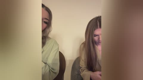 Media: A video of two young women with long hair, wearing light-colored robes, sitting side by side on a chair against a beige wall, their faces partially obscured by vertical light beams.