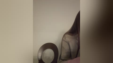 Media: Video of a woman with long brown hair, partially seen from the back, sitting on a bed. A metallic circular object is placed on the bed, creating a reflective surface. The background is a plain, beige wall.