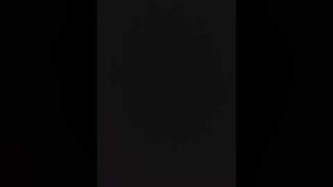 A black and white video of a man in a suit, standing against a dark background, his face obscured by shadow. The image is minimalist, emphasizing form and contrast.