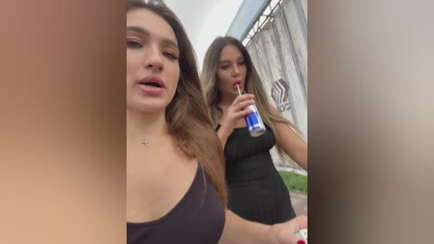 Media: A video featuring two young women with long hair and light skin tones, wearing black tops, walking outdoors. One holds a blue drink, while the other looks away.