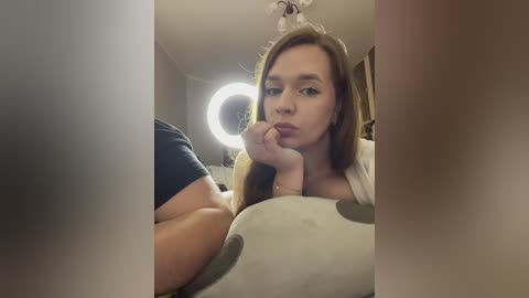 Media: A video of a young Caucasian woman with light brown hair, wearing a white top, lying on a bed with a circular light ring in the background, blurry.