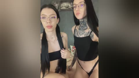 Video of two young women with pale skin, long black hair, and large glasses, one wearing a black bandeau top and panties, the other a black crop top, posing in a dimly lit room with tattooed necks.