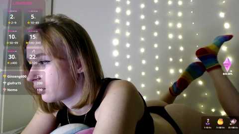 Video of a fair-skinned woman with shoulder-length brown hair, lying on her stomach in a black sports bra, wearing colorful striped socks, against a backdrop of string lights.