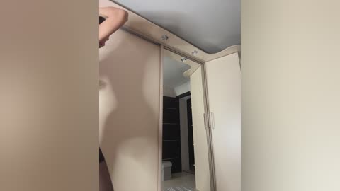 Video of a nude person with a small build, standing in front of a full-length mirror, reflected in a closet with white doors, in a dimly lit room with beige walls.