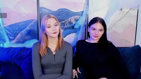 Video of two young women, one with long blonde hair in a grey top, the other with long black hair in a black top, seated on a dark couch against a backdrop of a scenic mountain landscape.