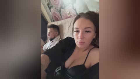 Media: Video of a young woman with dark hair and light skin, wearing a black bra, sitting next to a man with a beard and mustache, in a room with floral wallpaper.