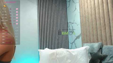Video of a modern bedroom with a white bed, gray and white striped wall, and beige curtains. A woman's arm is partially visible, holding a phone displaying health stats, on the left.