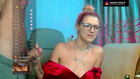 A video of a fair-skinned woman with red hair in a bun, wearing glasses and a red satin dress, seated in a living room with teal curtains and a grey couch, holding a phone.