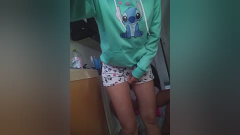 Video of a light-skinned woman in a green hoodie with a Stitch design, wearing patterned shorts. She stands in a cluttered kitchen with a sink, utensils, and a floral cup in the background.
