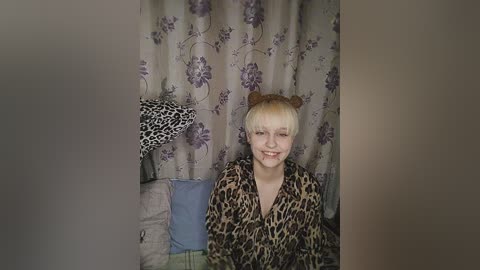 Media: Video of a blonde woman with short hair and brown bear ears, wearing a leopard print blouse, smiling, seated against floral-patterned curtains.