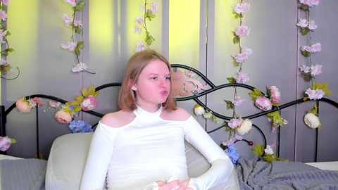 Video of a fair-skinned, blonde woman with shoulder-length hair in a white off-the-shoulder dress, seated on a bed with a grey headboard adorned with pink and white flowers. The background features a lavender curtain and more floral decor.