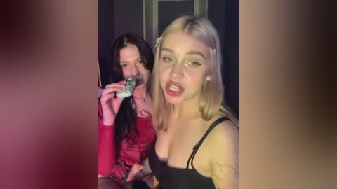 Media: Video of two young women in a dimly lit room. One, blonde, wearing a black top, leans forward. The other, with long black hair, holds a camera, dressed in red.
